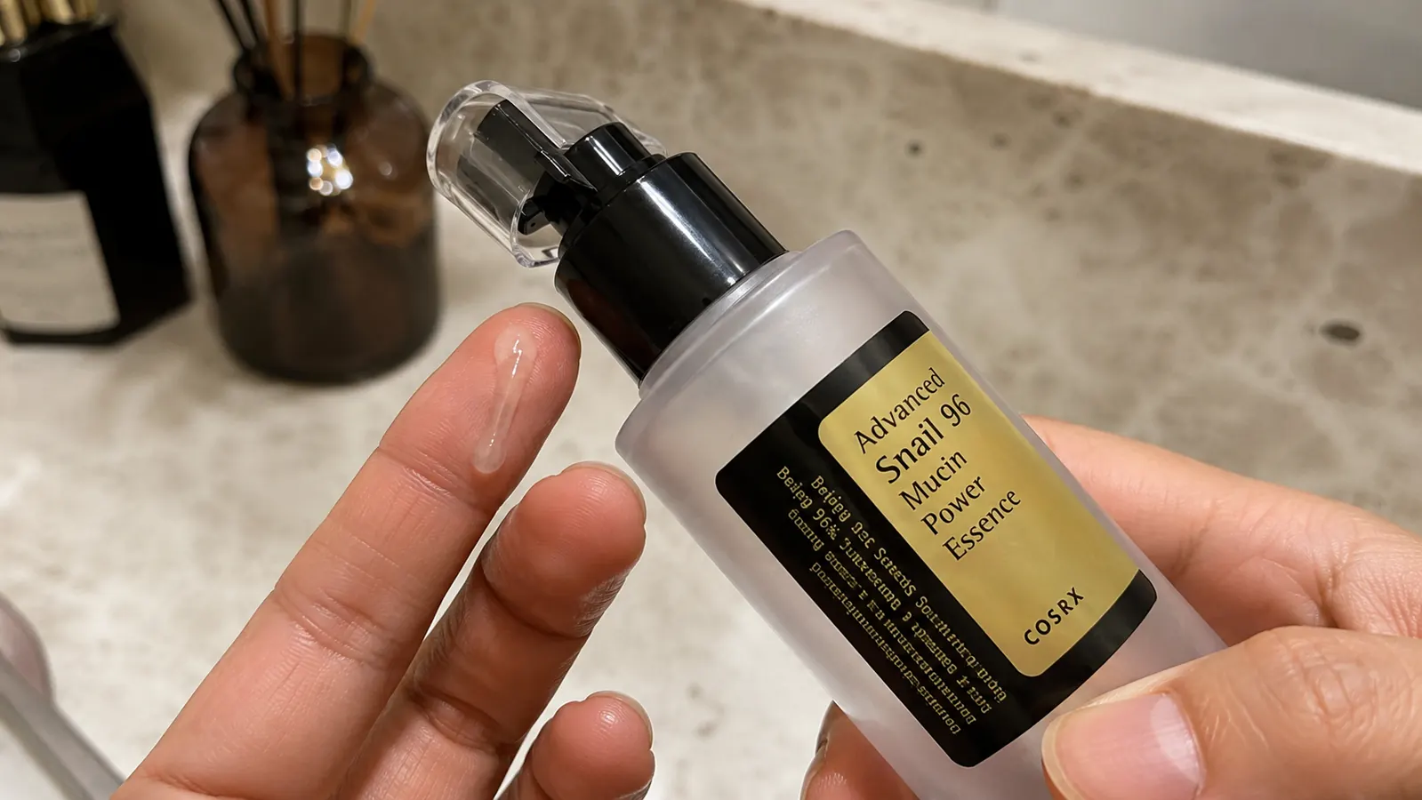 Hand holding the COSRX Snail Mucin essence bottle with a dab of clear serum on a fingertip