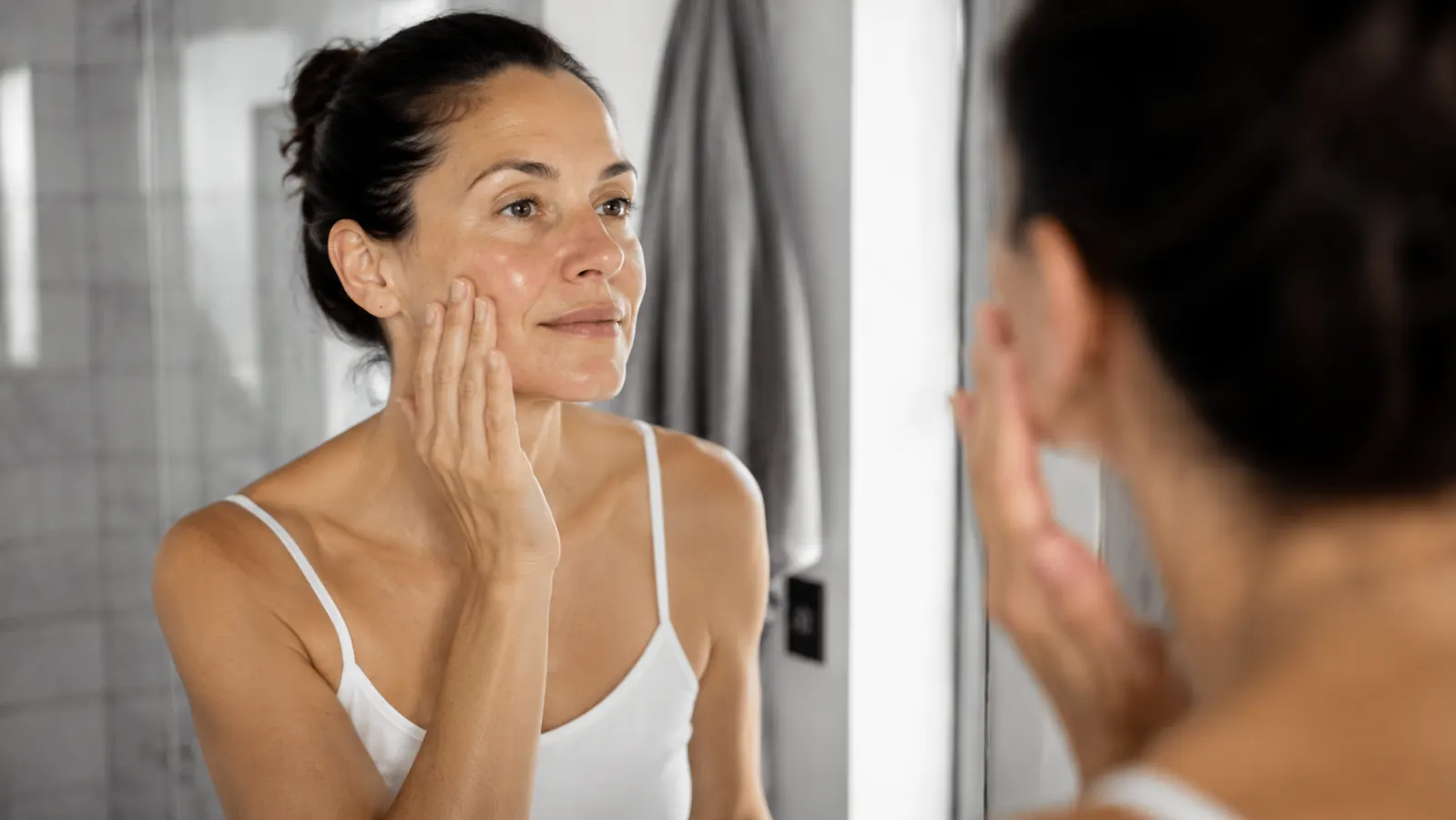 Person patting a snail mucin essence into their cheek at a bathroom sink in soft morning light