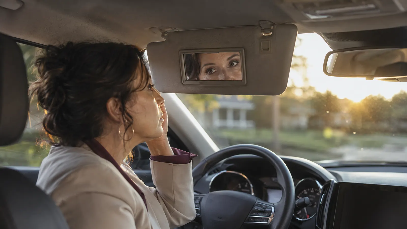 Woman in business casual sitting in a parked car at golden hour, checking her tired reflection in the visor mirror