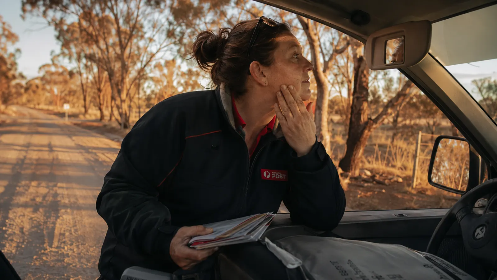 Woman in a postal delivery uniform standing beside her mail truck at dusk, touching her cheek, dry rural road in the background
