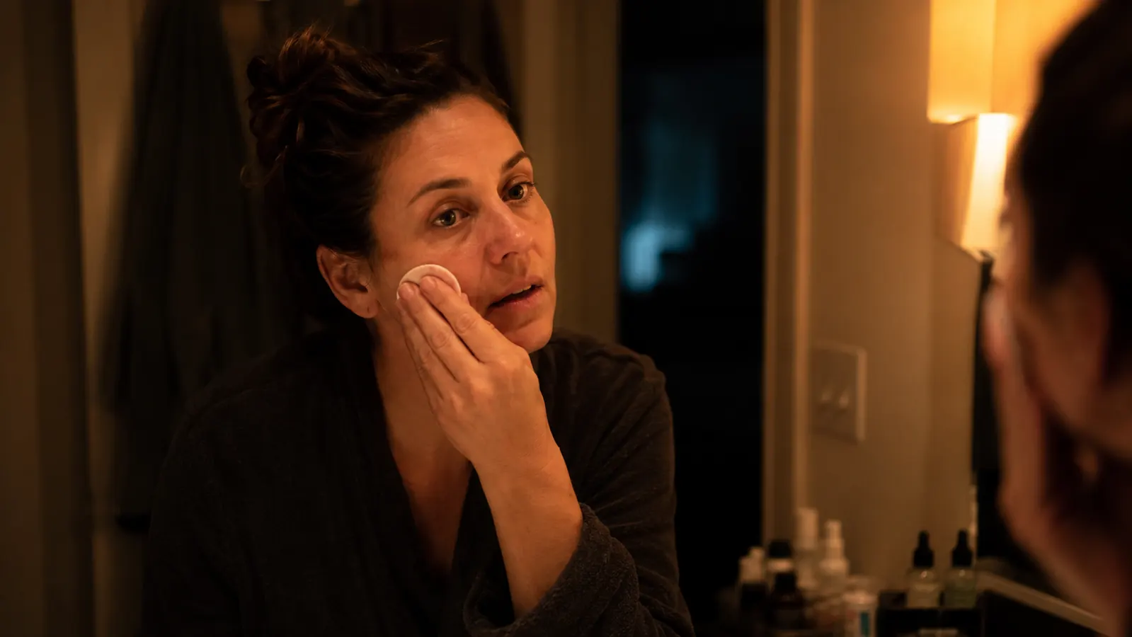 Woman applying a glycolic acid toner pad to her cheek at a bathroom sink in warm evening light