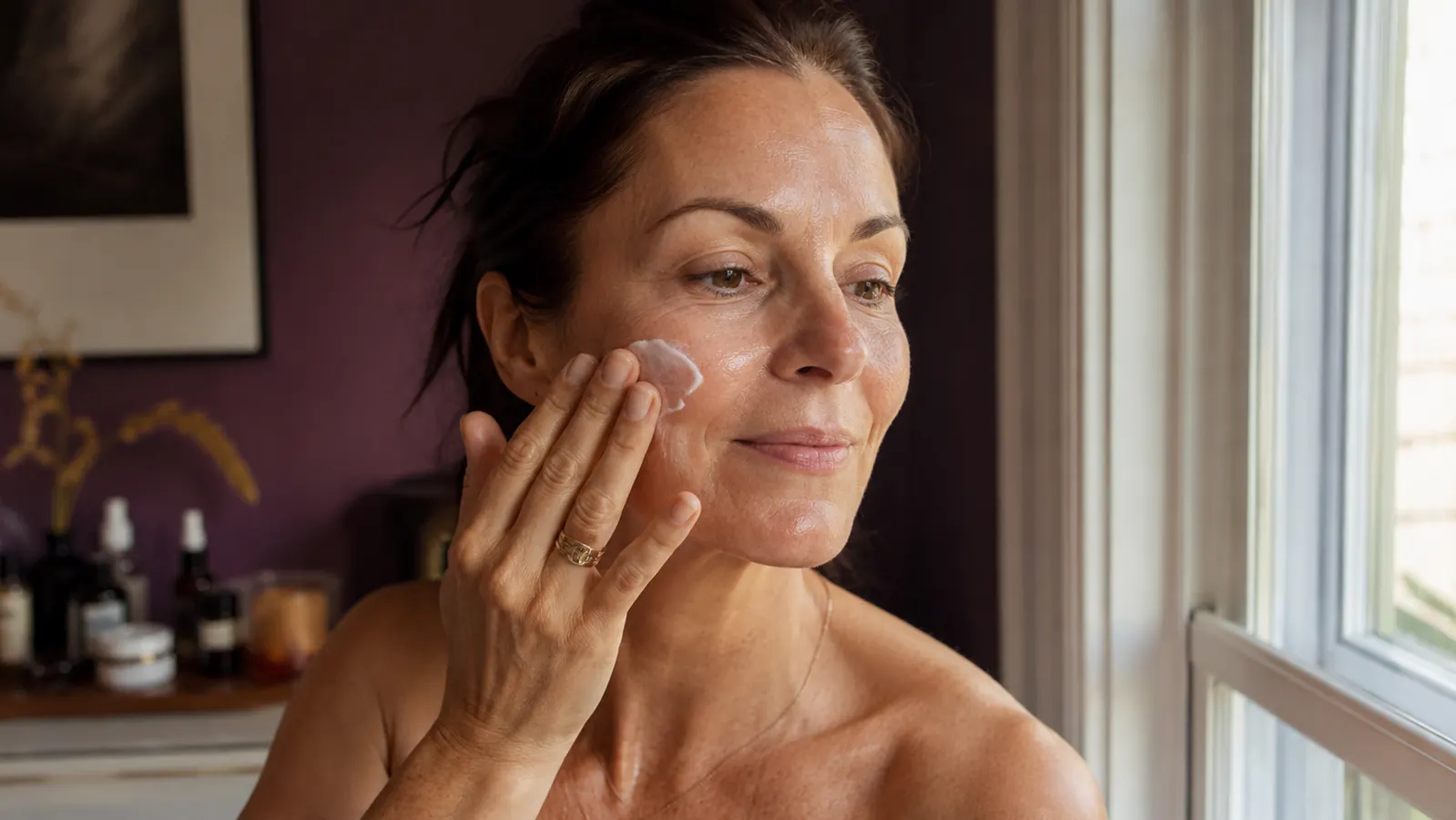 Woman with a fresh, dewy complexion applying moisturizer in daylight near a window, no makeup