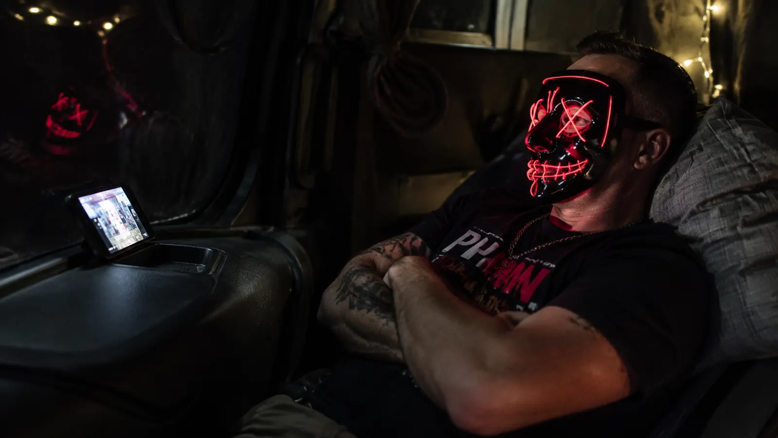 Man sitting in a truck sleeper cab at night wearing an illuminated red light therapy mask, phone propped against the window