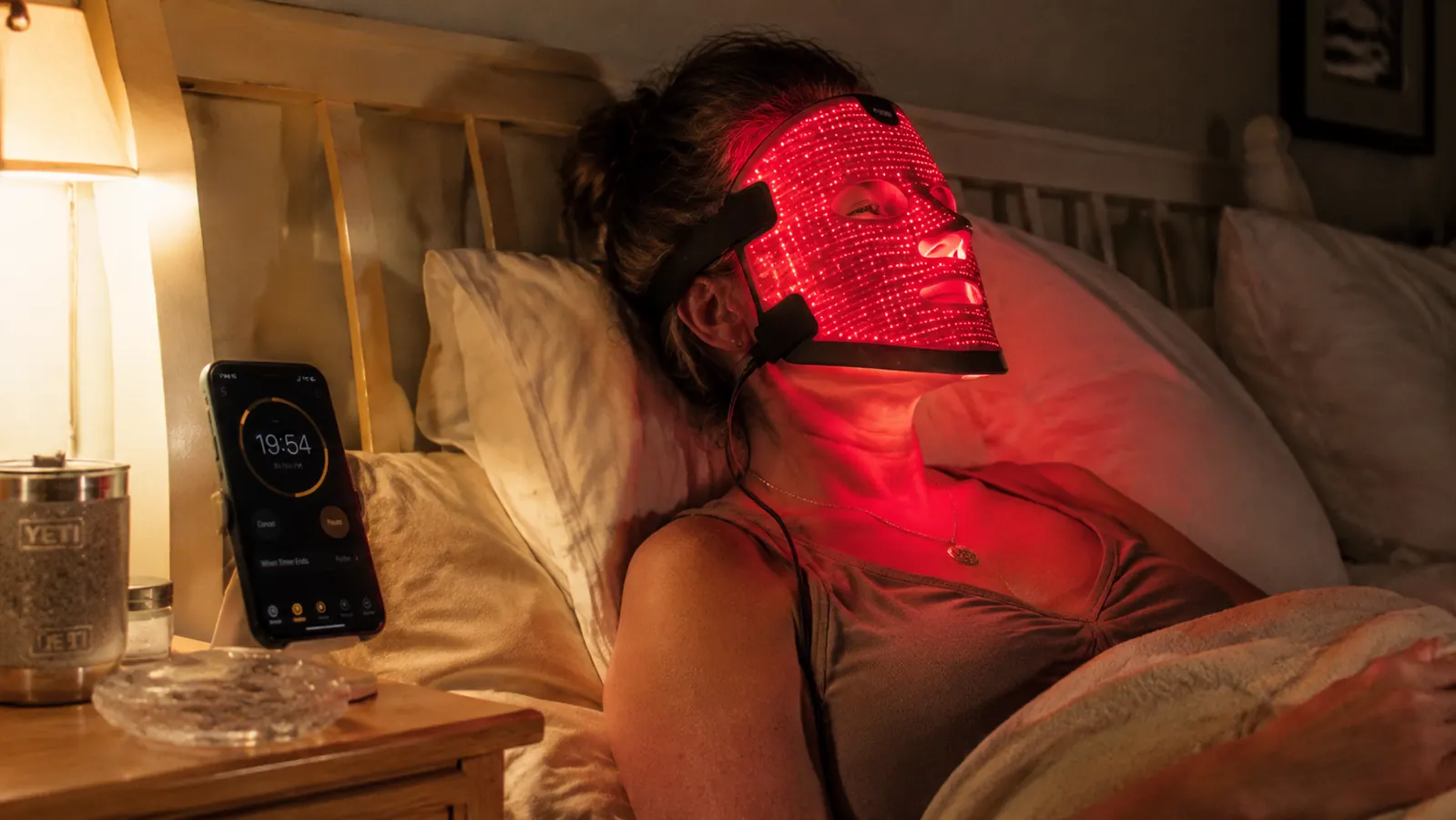Woman relaxing on her bed at night wearing a red light therapy mask, soft red glow lighting her face