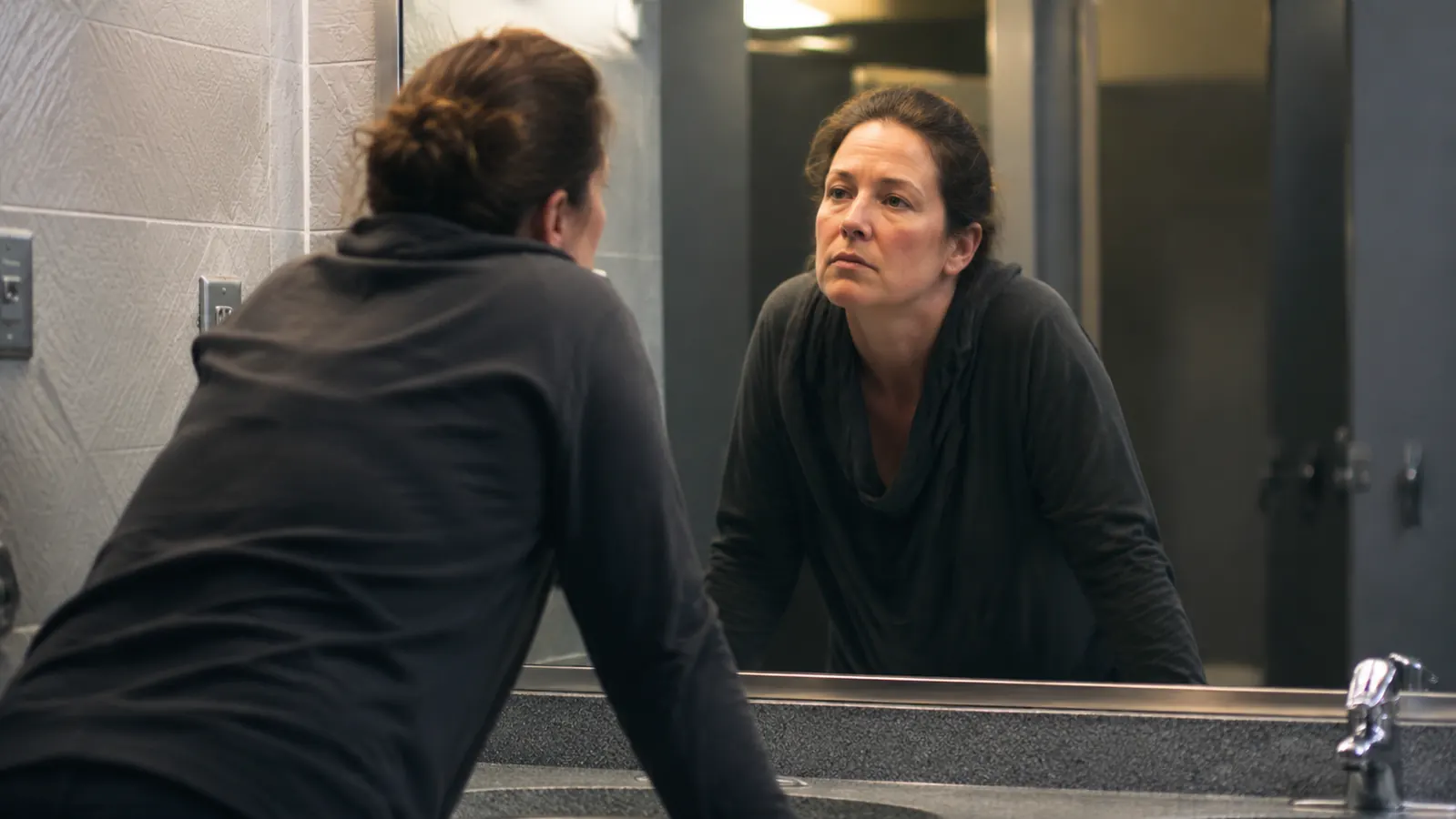 Woman checking her reflection in a truck stop mirror, morning light, tired but hopeful expression